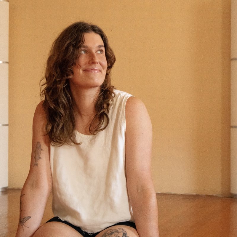 Haley Havelock, vinyasa yoga teacher at The Castro Room in San Francisco, seated on a wooden platform in a warm-toned studio, wearing a white tank top