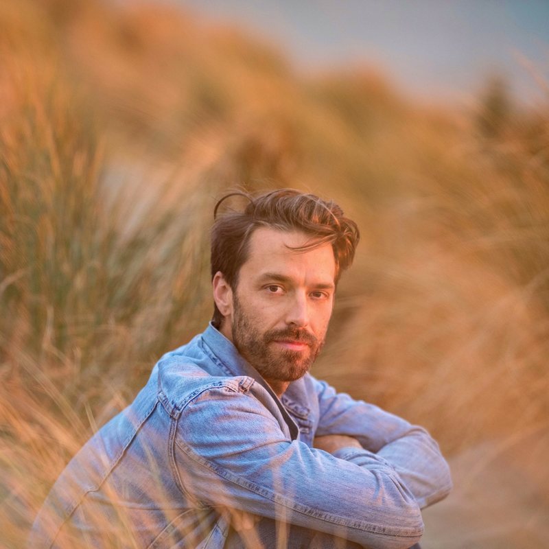 Jason Bowman, vinyasa yoga teacher at The Castro Room in San Francisco, seated among beach grass in golden evening light, wearing a denim jacket
