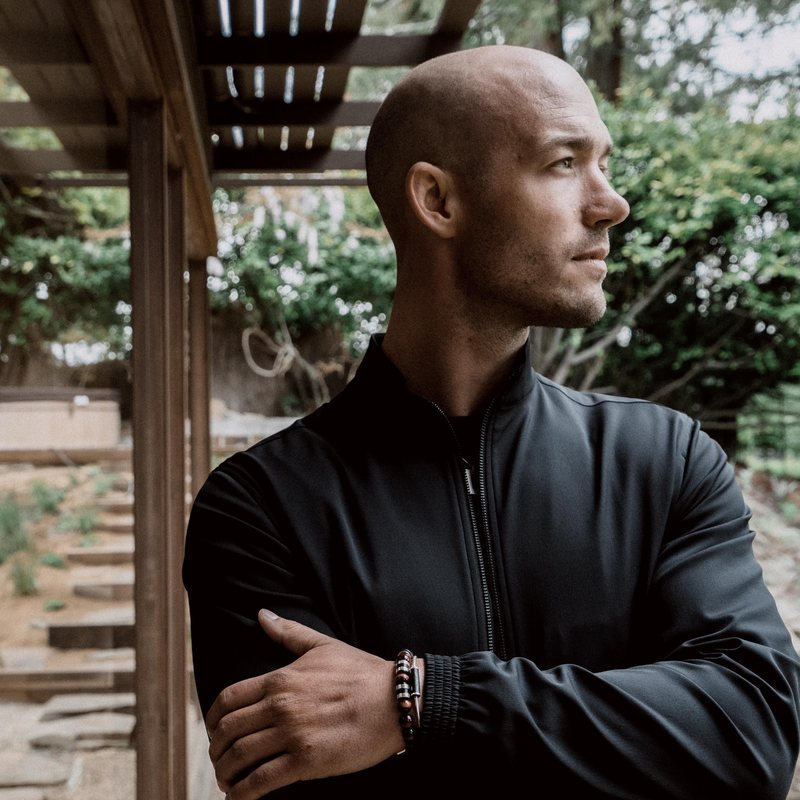 Peter Bartesch, vinyasa yoga teacher at The Castro Room in San Francisco, arms crossed, gazing to the side on a wooden porch surrounded by greenery