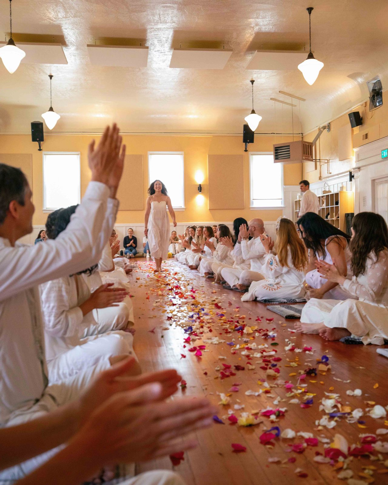 Yoga teacher training graduation: seated students in white applaud as a person walks a path of flower petals