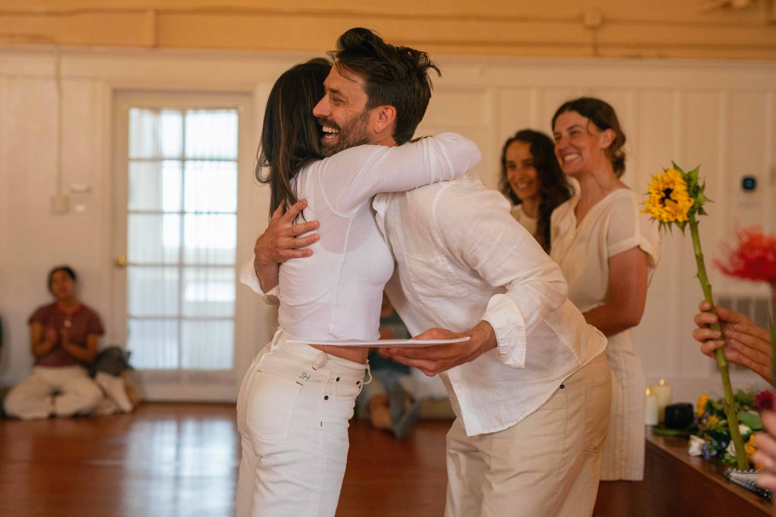 Two people in white embrace at the yoga teacher training graduation, with a sunflower in the foreground